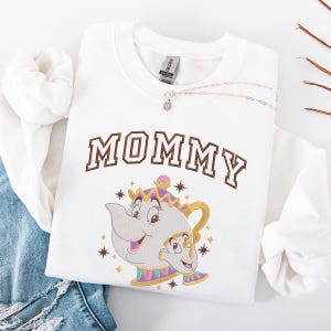 Può includere: Felpa bianca con la scritta "MOMMY" in lettere marroni. Sotto, un'immagine ricamata di Mrs. Bric e Chicco de La Bella e la Bestia. Una collana d'argento con un piccolo ciondolo è sulla felpa.