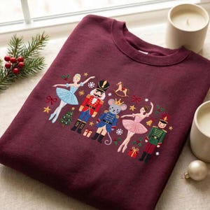 Könnte beinhalten: Ein bordeauxrotes Sweatshirt mit Rundhalsausschnitt und Stickereien von Nussknacker-Figuren, darunter eine Ballerina im blauen Kleid, ein Soldat, eine Maus in blauer Uniform, eine Ballerina im rosa Kleid und ein Soldat in grüner Uniform.