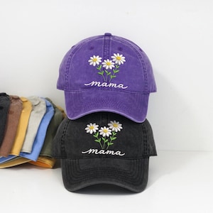 Può includere: Due cappellini da baseball, uno viola e uno nero, impilati l'uno sull'altro. Ogni cappellino presenta un ricamo di margherite bianche con steli verdi e la parola "mama" in corsivo. I cappellini sono realizzati in un tessuto morbido e usurato.