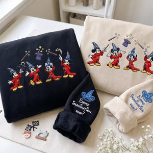 Peut inclure: Un sweat-shirt noir et un sweat-shirt beige, chacun orné de broderies représentant Mickey Mouse avec un chapeau de magicien bleu et une robe rouge. Les manches portent une broderie d'un chapeau bleu et le texte, Some imagination, huh?