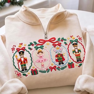 Könnte beinhalten: Ein cremefarbenes Sweatshirt mit kurzem Reißverschluss und einer Stickerei, die zwei Nussknacker, eine Ballerina und einen Mäusekönig zeigt, jeweils umrahmt von dekorativen Kränzen mit roten Schleifen, ergänzt durch kleine Geschenke, Zuckerstangen und Sterne.