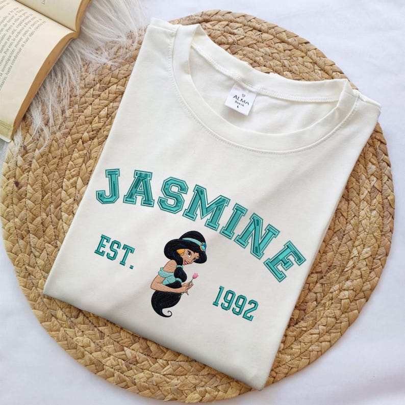 May include: A cream-colored short-sleeve t-shirt featuring the text JASMINE in teal block letters, the abbreviation EST., and the year 1992. A graphic of the character Jasmine holding a pink flower is centered below the text on the front.