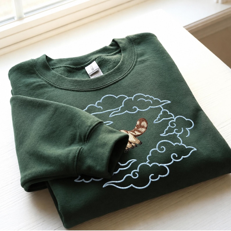 K&ouml;nnte beinhalten: Ein dunkelwaldgr&uuml;ner Rundhals-Pullover mit einer Stickerei, die ein braun-wei&szlig; gestreiftes Wesen zeigt, das durch hellblau gestickte Wolken fliegt. Das Kleidungsst&uuml;ck hat lange &Auml;rmel mit gerippten B&uuml;ndchen und einen gerippten Halsausschnitt.