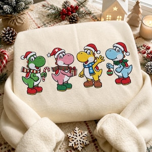 Peut inclure: Un sweat-shirt couleur crème avec une broderie représentant quatre personnages dinosaures colorés portant des bonnets de Père Noël. Les personnages vert, rose, jaune et bleu sont ornés d'objets de fête, dont une canne en sucre, un cadeau et une boule.