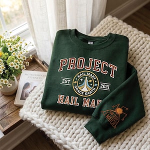 Può includere: Una felpa verde scuro con le parole "PROJECT HAIL MARY" in lettere bianche e marroni. La felpa presenta una grafica circolare con le parole "HAIL MARY GRACE EARTH" e l'anno "2021". La manica ha una grafica di un insetto e le parole "YOU SLEEP I WATCH".