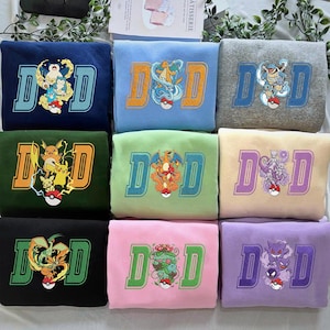 Può includere: Una collezione di nove felpe piegate in vari colori, ognuna con le lettere ricamate "D" e "D", un design di personaggi Pokémon e una Pokéball. I colori includono blu navy, azzurro, grigio, verde, verde chiaro, beige, nero, rosa e viola.