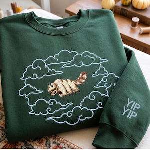 K&ouml;nnte beinhalten: Ein dunkelgr&uuml;ner Rundhals-Pullover mit einer Stickerei eines beigen und braunen fliegenden Bisons, umgeben von hellblau gestickten Wolken. Auf dem rechten &Auml;rmel befindet sich der gestickte Schriftzug YIP YIP in hellblauem Garn.