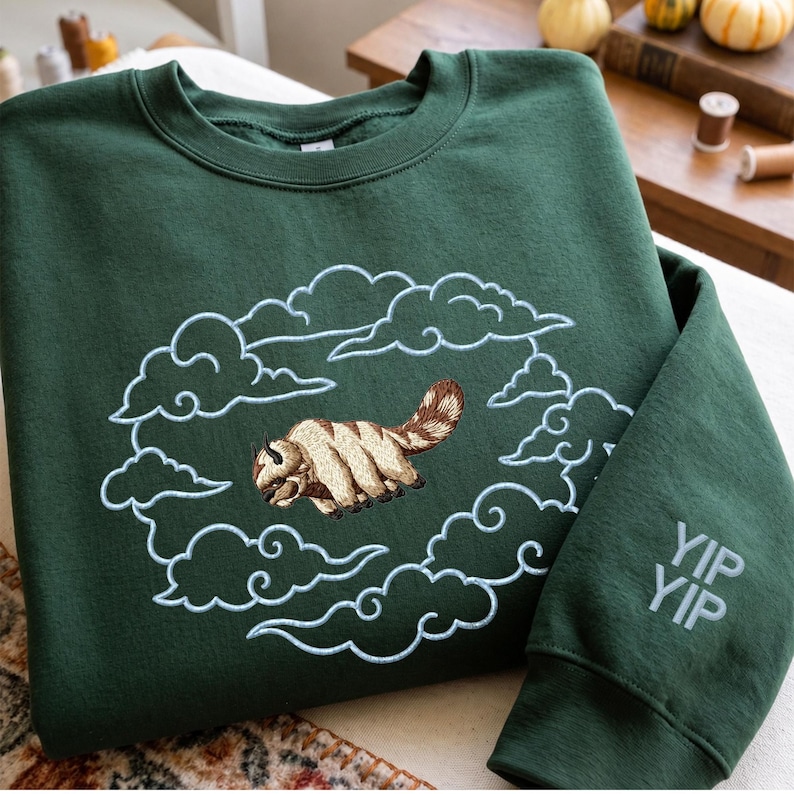 K&ouml;nnte beinhalten: Ein dunkelgr&uuml;ner Rundhals-Pullover mit einer Stickerei eines beigen und braunen fliegenden Bisons, umgeben von hellblau gestickten Wolken. Auf dem rechten &Auml;rmel befindet sich der gestickte Schriftzug YIP YIP in hellblauem Garn.