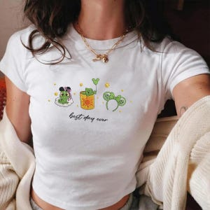 Peut inclure: Un t-shirt blanc à manches courtes orné d'un motif représentant un caméléon vert dans trois positions différentes, avec des oreilles de souris, dans une tasse à motif soleil et avec un serre-tête. Le texte "best day ever" est écrit en noir.