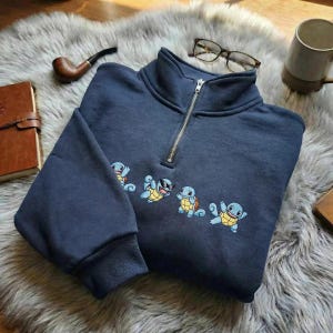 Peut inclure: Un sweat-shirt bleu marine à fermeture éclair quart de tour, orné d'un motif brodé représentant quatre personnages de tortues bleu clair dans diverses poses. Le vêtement possède un col montant avec une fermeture éclair argentée.
