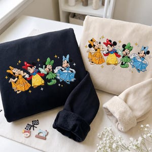 Può includere: Una felpa nera e una felpa beige, ciascuna caratterizzata da una grafica ricamata di quattro personaggi di Minnie Mouse vestiti con colorati abiti da principessa, accentuati da piccole stelle ricamate attorno alle figure.