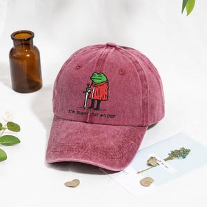 Könnte beinhalten: Weinrote Baseballkappe mit Used-Look. Auf der Kappe ist ein gestickter Cartoon-Frosch mit rotem Mantel und Schwert abgebildet. Der Text "I'M BURNT OUT M'LORD" ist darunter gestickt.