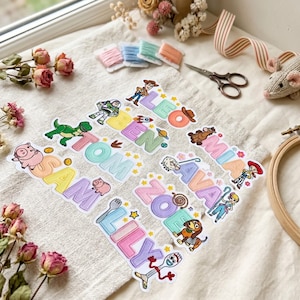 Op de afbeelding: Een verzameling geborduurde patches met namen zoals Leo, Mia, Ben, Tom, Ava, Sam, Zoe en Lily, vergezeld door kleurrijke Toy Story stripfiguren, waaronder Woody, Buzz Lightyear, Rex, Hamm, Jessie, Bo Peep, Slinky Dog en Forky.