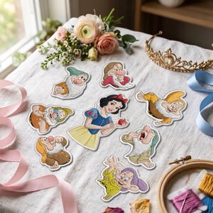 May include: A collection of embroidered patches featuring Snow White holding a red apple, surrounded by the seven dwarfs. The patches are arranged on a white fabric surface alongside a gold tiara, pink ribbons, and a wooden embroidery hoop.