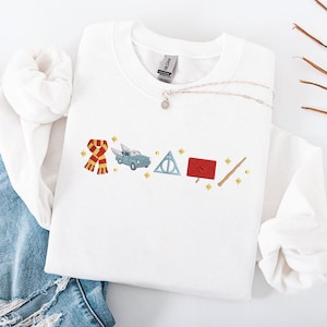Op de afbeelding: Witte crewneck sweatshirt met geborduurde Harry Potter-thema ontwerpen. Voorzien van een rood-gouden sjaal, een blauwe auto met vleugels, een Deathly Hallows symbool, een rode envelop en een toverstaf. Een zilveren ketting met een kleine hanger is ook zichtbaar.