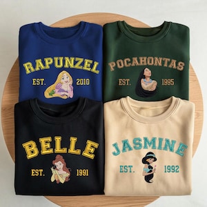 May include: Four crewneck sweatshirts in blue, dark green, black, and beige. Each features a Disney princess graphic with their name in varsity-style lettering and an establishment year: Rapunzel 2010, Pocahontas 1995, Belle 1991, and Jasmine 1992.