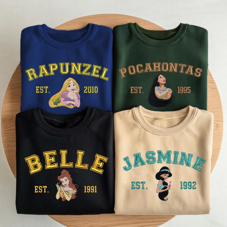 May include: Four crewneck sweatshirts in blue, dark green, black, and beige. Each features a Disney princess graphic with their name in varsity-style lettering and an establishment year: Rapunzel 2010, Pocahontas 1995, Belle 1991, and Jasmine 1992.