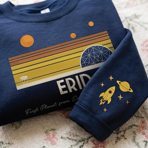 Puede incluir: Sudadera azul marino con un diseño de temática espacial. La parte delantera presenta un gráfico retro de puesta de sol con la palabra "ERID" y una ilustración de un planeta. La manga muestra un cohete amarillo, un planeta y estrellas.