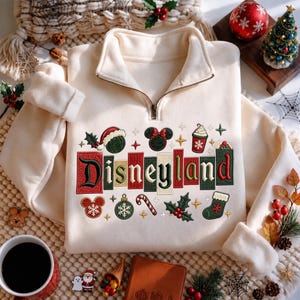 Può includere: Una felpa color crema con mezza zip e un ricamo della parola Disneyland in lettere colorate. Il design include icone natalizie come un cappello da Babbo Natale, orecchie di Topolino, una tazza di caffè, un bastoncino di zucchero e dell'agrifoglio.