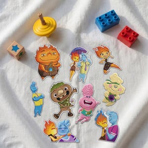 May include: A collection of colorful cartoon character stickers featuring various figures with fire, water, and earth-like traits, accompanied by a yellow spinning top, a wooden block with a blue star, and two plastic building bricks in blue and red.