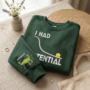 Könnte beinhalten: Ein dunkelgrünes Sweatshirt mit dem weißen Text "I HAD POTENTIAL". Der Ärmel zeigt einen Cartoon-Frosch mit dem Wort "amaze!". Das Sweatshirt ist ein lässiges Kleidungsstück.