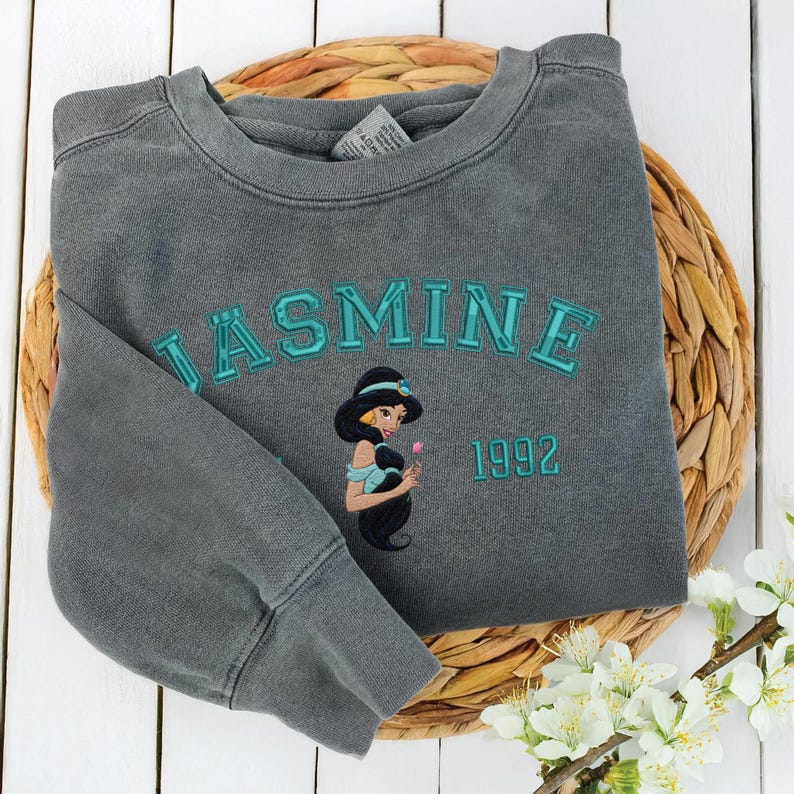 May include: A charcoal gray crewneck sweatshirt featuring the embroidered text JASMINE in teal block letters, a graphic of the character Jasmine, and the year 1992 in teal. The garment has long sleeves and a ribbed collar, cuffs, and hem.