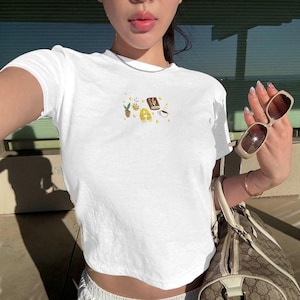 May include: White crop top with a small, colorful graphic of a book, coffee cup, and other objects. The shirt is short-sleeved and has a crew neck. The person is holding sunglasses and a patterned bag.