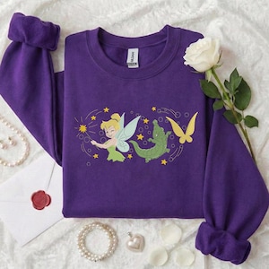Op de afbeelding: Een paarse sweatshirt met een ronde hals en lange mouwen. De voorkant heeft een geborduurd ontwerp van een fee, een groene alligator en een gele vlinder, met sterren. De sweatshirt is gemaakt van een zacht materiaal.