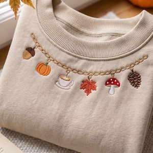 Puede incluir: Una sudadera beige de cuello redondo con un diseño de cadena dorada bordada en el pecho. La cadena está adornada con pequeños dijes bordados de colores, que incluyen una bellota, una calabaza, una taza de café, una hoja de arce, una seta roja y una piña.