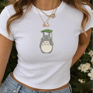 Könnte beinhalten: Ein weißes kurzärmeliges T-Shirt mit einer mittigen Grafik einer grauen, runden Zeichentrickfigur mit Schnurrhaaren und einem cremefarbenen Bauch, die ein großes grünes Blatt über dem Kopf hält. Das Shirt hat einen Rundhalsausschnitt.