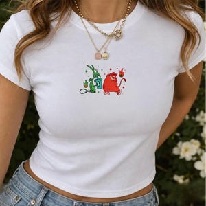 Puede incluir: Una camiseta blanca de manga corta con un pequeño estampado gráfico en el centro. El diseño muestra una criatura verde sosteniendo una taza azul y una criatura roja con cola, ambas rodeadas de pequeños iconos de llamas y rombos rojos.