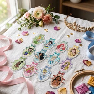 Op de afbeelding: Een collectie van twaalf ovaalvormige geborduurde patches met diverse geanimeerde personages, elk omlijst door een decoratieve rand en een klein gekleurd lintstrikje. De patches liggen op een witte stoffen ondergrond met bloemen en een gouden tiara.