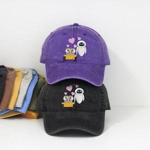 Può includere: Due cappellini da baseball impilati, uno viola e uno nero. Ogni cappellino presenta un disegno ricamato di un robot giallo, un robot bianco e cuori rosa. I cappellini hanno una visiera curva e un cinturino regolabile.