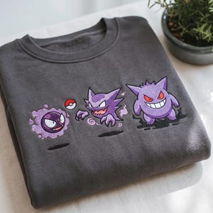 Op de afbeelding: Een antracietgrijze sweater met ronde hals en een geborduurde afbeelding van drie paarse spookachtige wezens op een rij. Het ontwerp bevat een klein rood-wit bal-icoon en gedetailleerd stikwerk van de figuren met rode ogen en scherpe tanden.