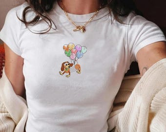 May include: White, short-sleeved t-shirt featuring a cartoon dog holding colorful balloons. The dog is brown, tan, and orange, with a green collar. The balloons are various pastel colors. The shirt is plain and has a crew neck.