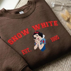 May include: A dark brown crewneck sweatshirt featuring an embroidered graphic of the character Snow White. The text "SNOW WHITE" is arched above the character, with "EST." on the left and "1995" on the right, all stitched in bright red thread.