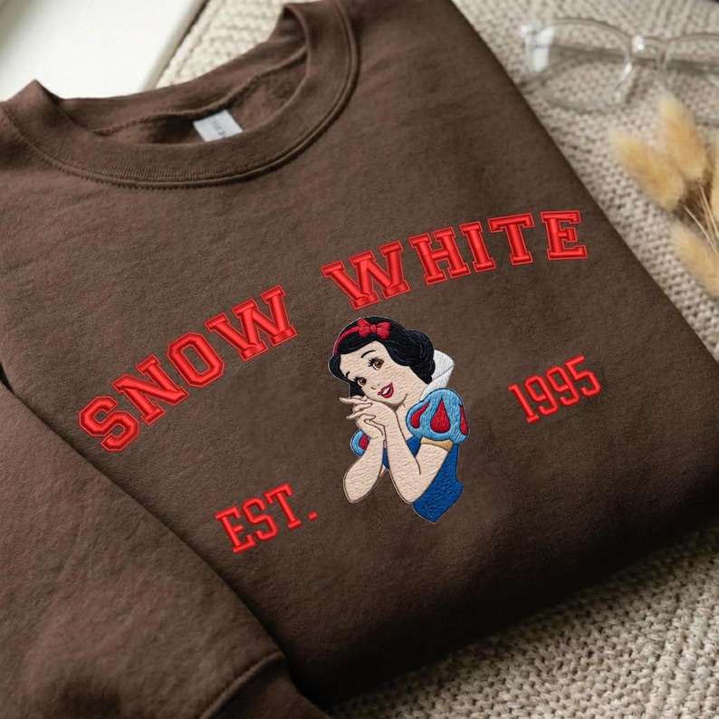 May include: A dark brown crewneck sweatshirt featuring an embroidered graphic of the character Snow White. The text "SNOW WHITE" is arched above the character, with "EST." on the left and "1995" on the right, all stitched in bright red thread.