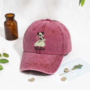 May include: A maroon baseball cap featuring an embroidered Minnie Mouse design in a bridal gown. The word "Bride" is embroidered below the character. The cap has a curved brim and a faded, vintage look.
