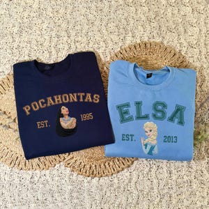 May include: A navy blue sweatshirt featuring an embroidered Pocahontas graphic with the text "POCAHONTAS EST. 1995" and a light blue sweatshirt featuring an embroidered Elsa graphic with the text "ELSA EST. 2013" in a collegiate-style font.