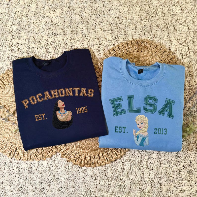 May include: A navy blue sweatshirt featuring an embroidered Pocahontas graphic with the text "POCAHONTAS EST. 1995" and a light blue sweatshirt featuring an embroidered Elsa graphic with the text "ELSA EST. 2013" in a collegiate-style font.