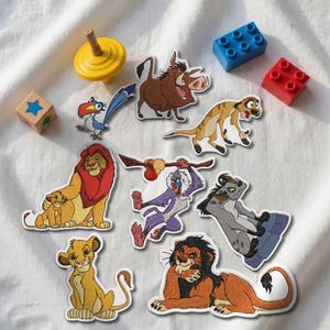 Puede incluir: Una colección de parches bordados con personajes de El Rey León, incluyendo a Simba, Mufasa, Scar, Timon, Pumbaa, Zazu, Rafiki y una hiena, dispuestos junto a una peonza amarilla, un bloque azul, un bloque rojo y un cubo de madera.