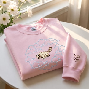 K&ouml;nnte beinhalten: Ein hellrosa Sweatshirt mit Rundhalsausschnitt und einer Stickerei eines fliegenden Bisons, umgeben von hellblau gestickten Wolken. Auf dem &Auml;rmel befindet sich der braun gestickte Text YIP YIP. Das Kleidungsst&uuml;ck hat gerippte B&uuml;ndchen.