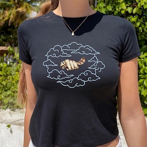 K&ouml;nnte beinhalten: Ein schwarzes Kurzarm-T-Shirt mit einer Stickerei, die ein beiges, fellartiges, sechsbeiniges Wesen mit gestreiftem Schwanz zeigt, das innerhalb einer kreisf&ouml;rmigen Umrandung aus hellblauen Wolken fliegt. Das Motiv ist mittig auf der Brust platziert.