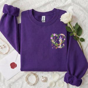 Könnte beinhalten: Ein lila Sweatshirt mit einem herzförmigen Blumendesign mit einem Turm und dem Buchstaben "R". Das Sweatshirt hat lange Ärmel mit gefalteten Bündchen. Eine weiße Rose und Schmuck sind um das Sweatshirt angeordnet.