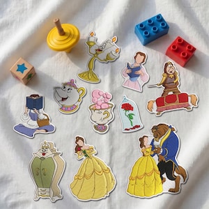 Può includere: Una collezione di toppe ricamate con personaggi e oggetti de La Bella e la Bestia, tra cui Belle, la Bestia, Lumière, Tockins, Mrs. Bric, Chicco, la rosa incantata e un armadio, insieme a una trottola gialla e dei mattoncini da costruzione.