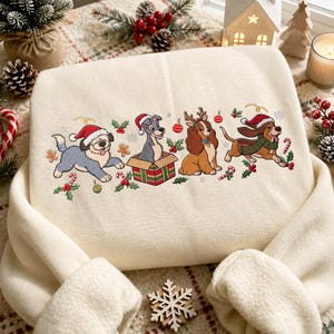 Può includere: Una felpa color crema con un ricamo raffigurante quattro cani dei cartoni animati tratti da Lilli e il Vagabondo. I cani indossano accessori natalizi come cappelli da Babbo Natale, corna di renna e sciarpe, circondati da agrifoglio, bastoncini.