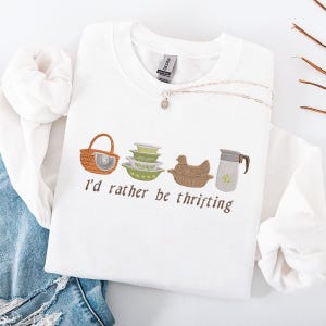 Może przedstawiać: Biała bluza z okrągłym dekoltem z haftowanymi obrazami koszyka, misek, kury i dzbanka. Pod spodem wyhaftowany jest napis "I'd rather be thrifting". Widoczny jest również srebrny naszyjnik z małą zawieszką.