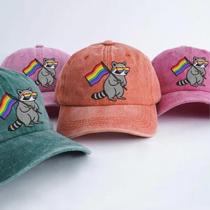 Può includere: Una collezione di cappellini da baseball in vari colori, tra cui verde acqua, rosa e arancione. Ogni cappellino presenta un procione ricamato con occhiali da sole e una bandiera arcobaleno. I cappellini sono realizzati in un tessuto morbido e strutturato.