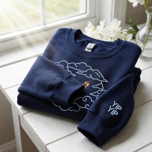 K&ouml;nnte beinhalten: Ein marineblaues Sweatshirt mit Rundhalsausschnitt, das auf der Brust ein hellblau gesticktes Wolkenmotiv und am linken &Auml;rmelb&uuml;ndchen den in Hellblau gestickten Schriftzug YIP YIP aufweist. Das Kleidungsst&uuml;ck hat gerippte B&uuml;ndchen.