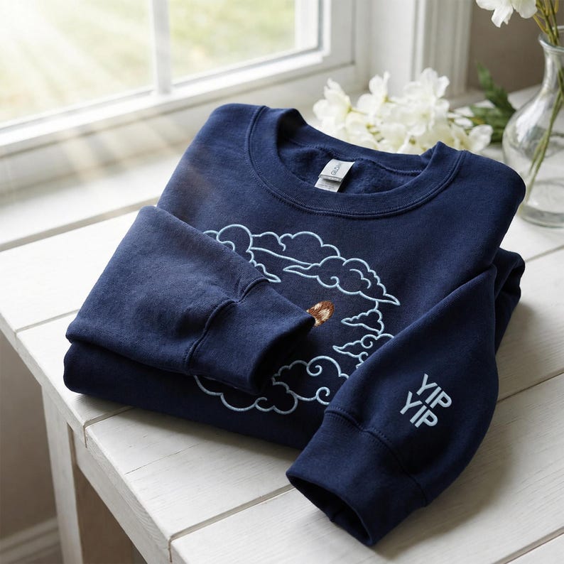 K&ouml;nnte beinhalten: Ein marineblaues Sweatshirt mit Rundhalsausschnitt, das auf der Brust ein hellblau gesticktes Wolkenmotiv und am linken &Auml;rmelb&uuml;ndchen den in Hellblau gestickten Schriftzug YIP YIP aufweist. Das Kleidungsst&uuml;ck hat gerippte B&uuml;ndchen.